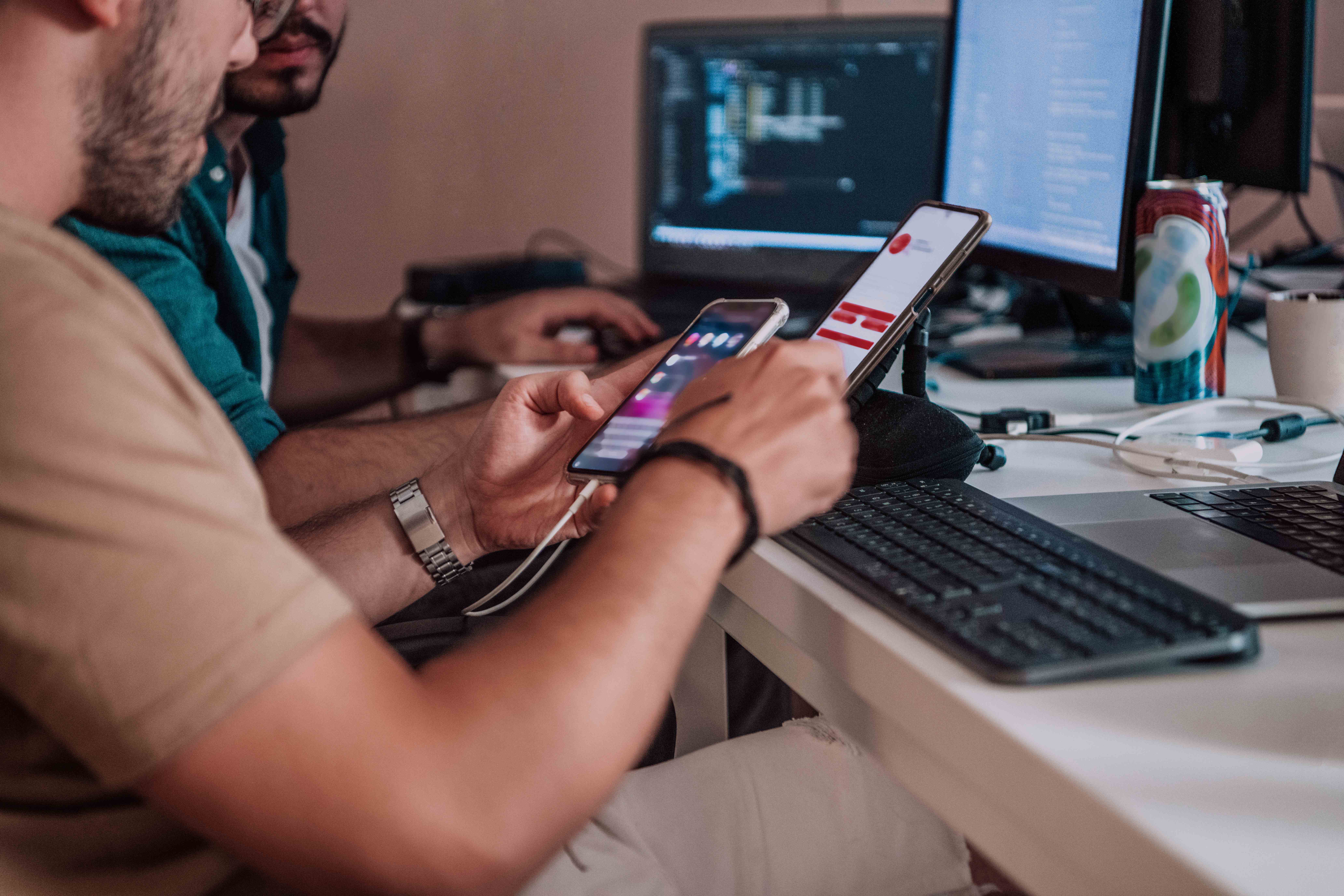 A developer sitting at a desk using a smartphone while working on a computer with multiple monitors displaying code in the background