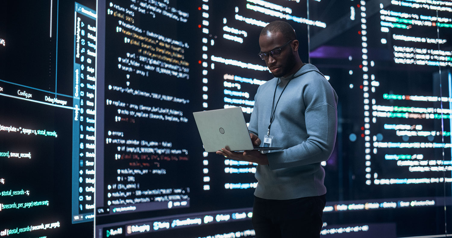 Developer reviewing code on a laptop surrounded by large screens displaying software scripts