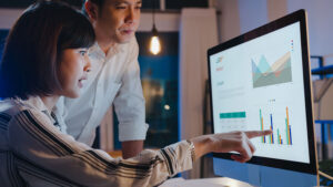 Two professionals reviewing charts and graphs on a monitor in a dimly lit office