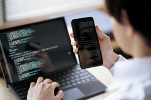 Developer testing mobile app on smartphone alongside laptop with code for cross-platform compatibility testing