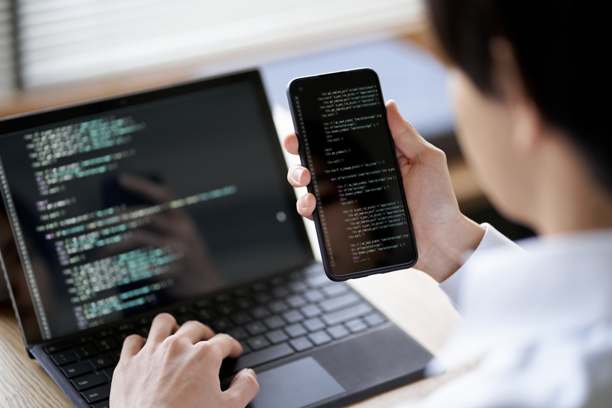 Developer testing mobile app on smartphone alongside laptop with code for cross-platform compatibility testing