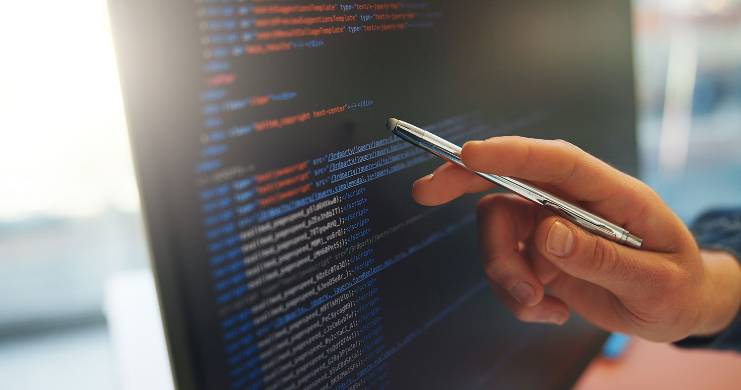 Hand holding a pen pointing at colorful code on a dark monitor screen