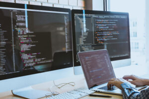 Developer coding on a laptop with two monitors showing syntax-highlighted code