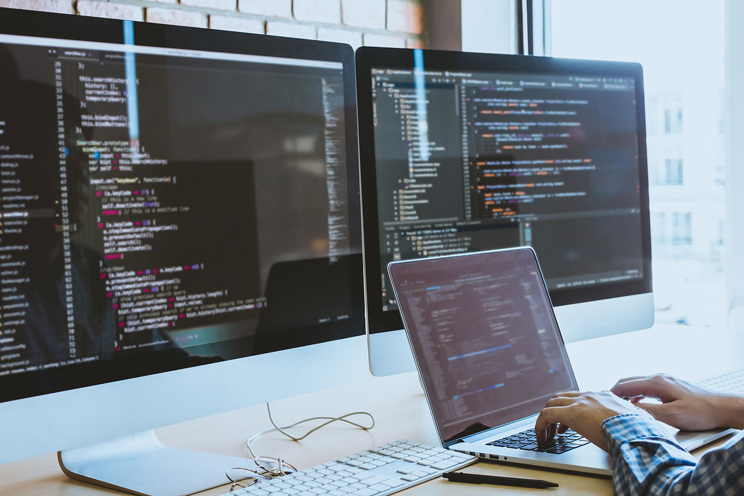 Developer coding on a laptop with two monitors showing syntax-highlighted code